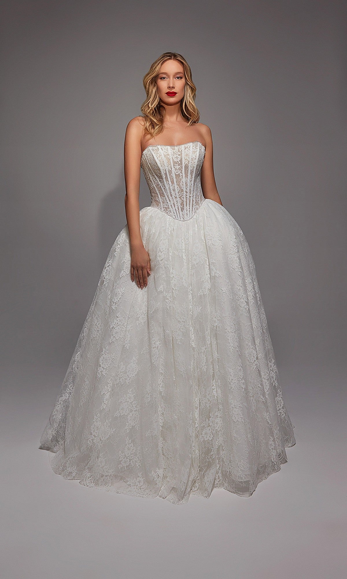 A woman poses against a plain gray background, wearing the Alyce 7149 Long White Dress featuring a strapless fitted bodice and a full lace skirt.
